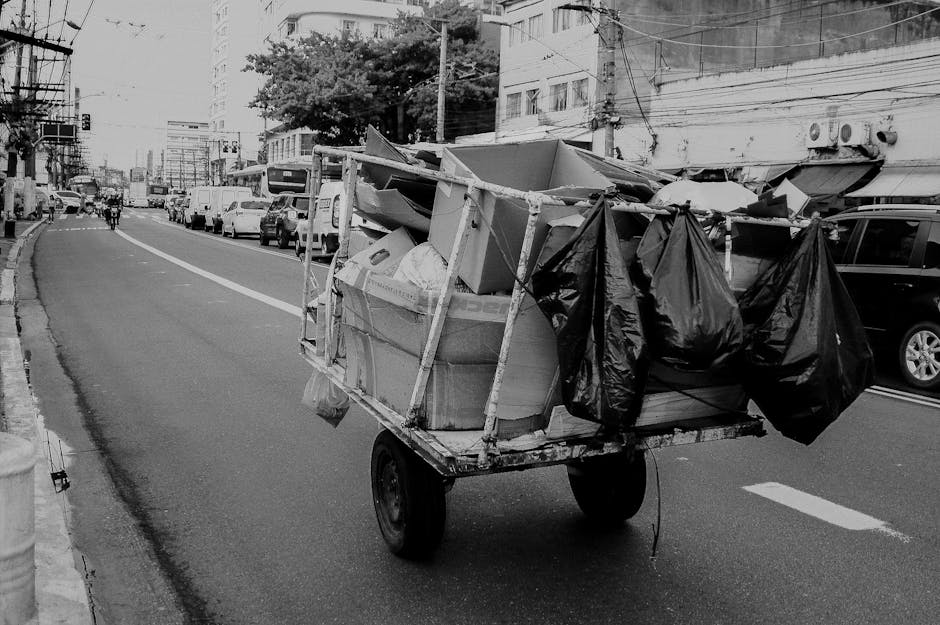 A small, manually operated cart with two wheels is loaded with a variety of discarded items on a city street. The cargo includes cardboard boxes, some of which are partially crushed, along with multip