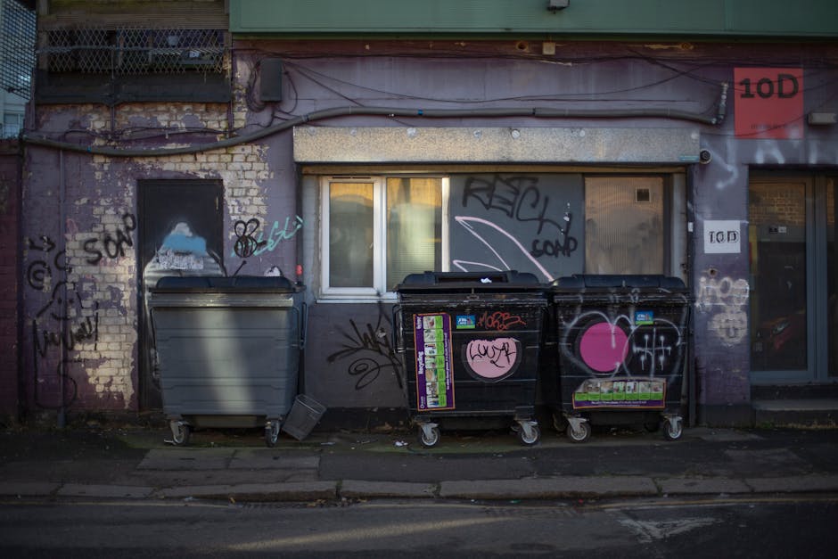 A small, manually operated cart with two wheels is loaded with a variety of discarded items on a city street. The cargo includes cardboard boxes, some of which are partially crushed, along with multip
