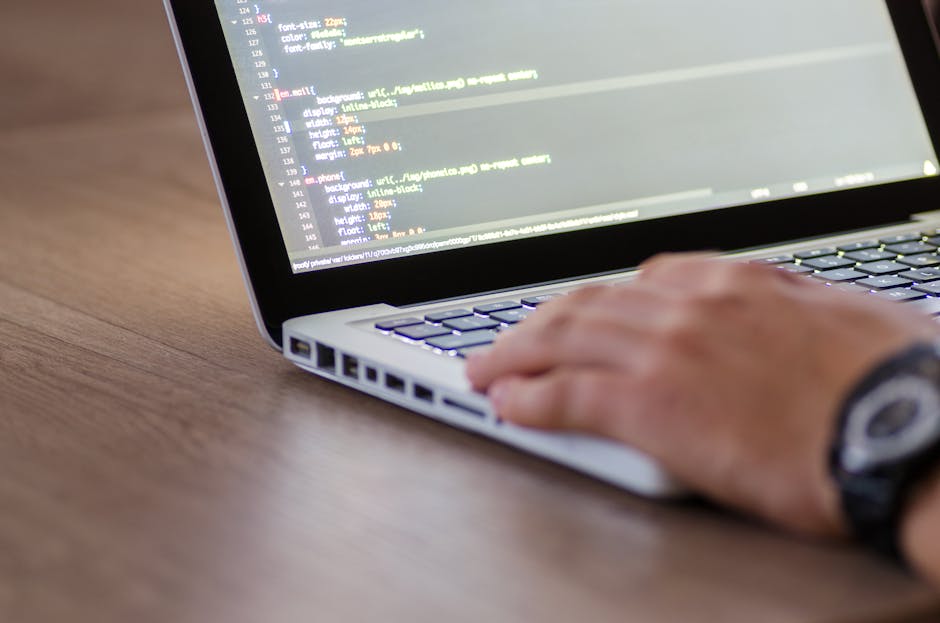 A close-up view of a person's right hand typing on a silver laptop keyboard, which is positioned on a wooden surface with a natural grain and warm brown tone. Part of the laptop's black screen is visi
